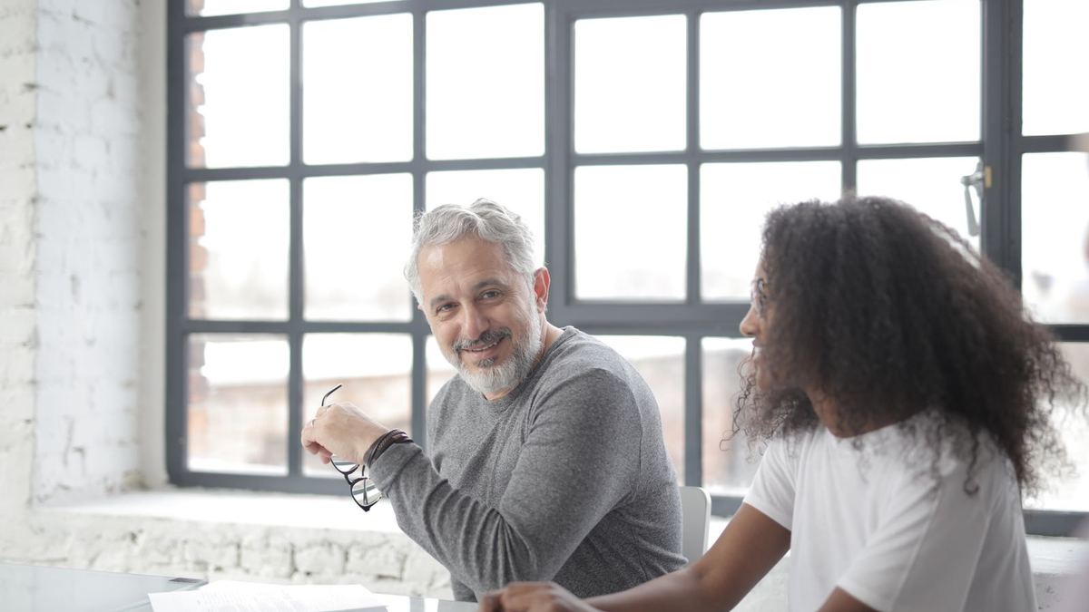 A gray-haired man and a young woman sit across a table by a large window in conversation, the woman listening as he talks.
