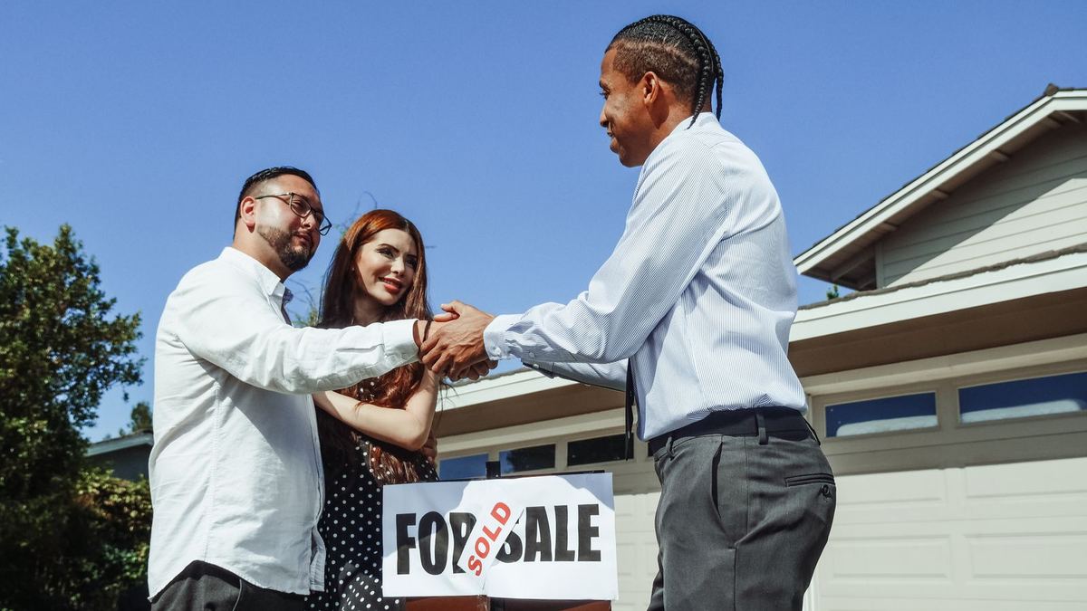 A real estate agent in a light blue shirt shakes hands with a smiling couple on the lawn in front of a house, with a red-and-white For Sale sign marked Sold between them.