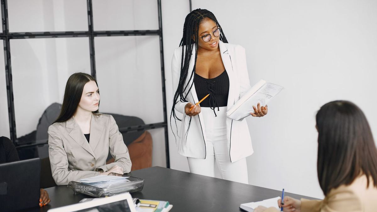 A woman in a white blazer stands holding papers and presents to three seated colleagues in a loft-style conference room.
