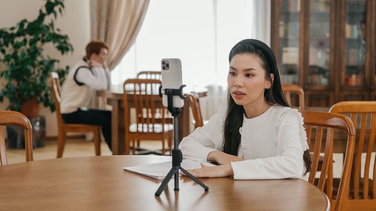 A woman sits at a dining table speaking to a smartphone mounted on a small tripod while recording a video.