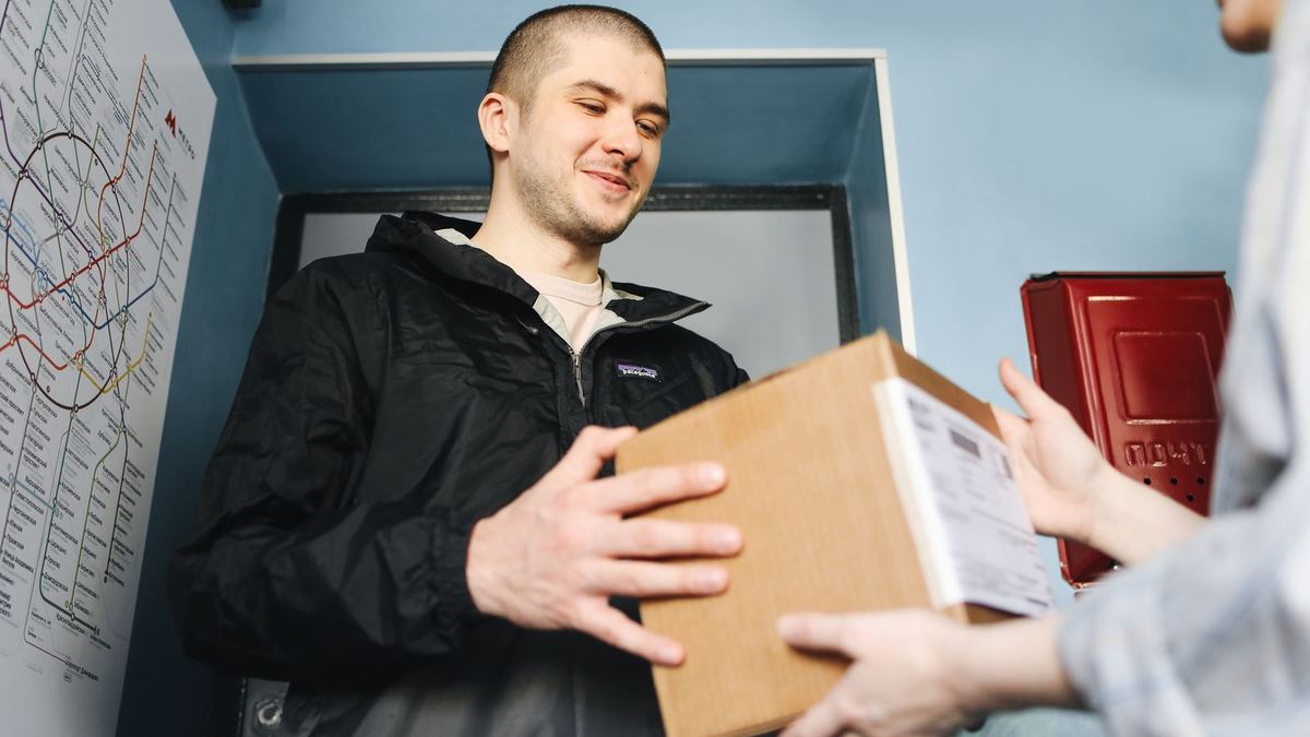 A delivery courier in a dark jacket hands a cardboard parcel to a customer at an apartment doorway.