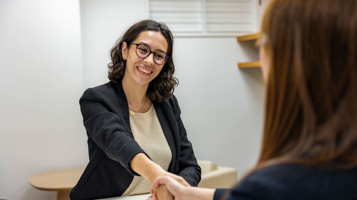 A smiling woman in a dark blazer shakes hands across a table with an interviewer in an office.